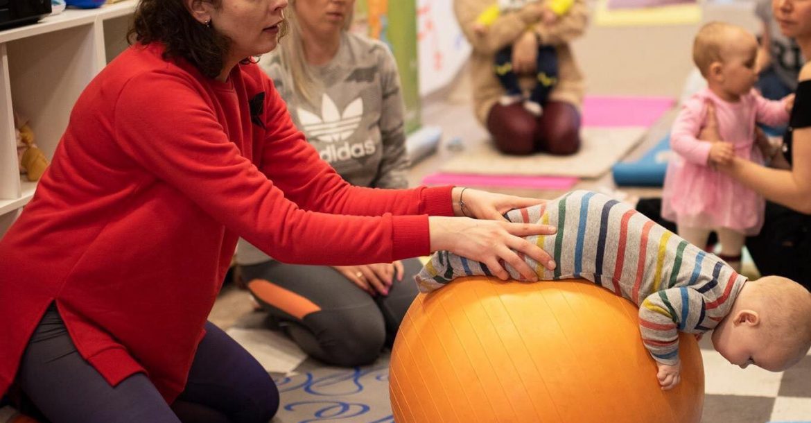 Kineziterapiautė mankština kūdikį ant oranžinio kamuolio
